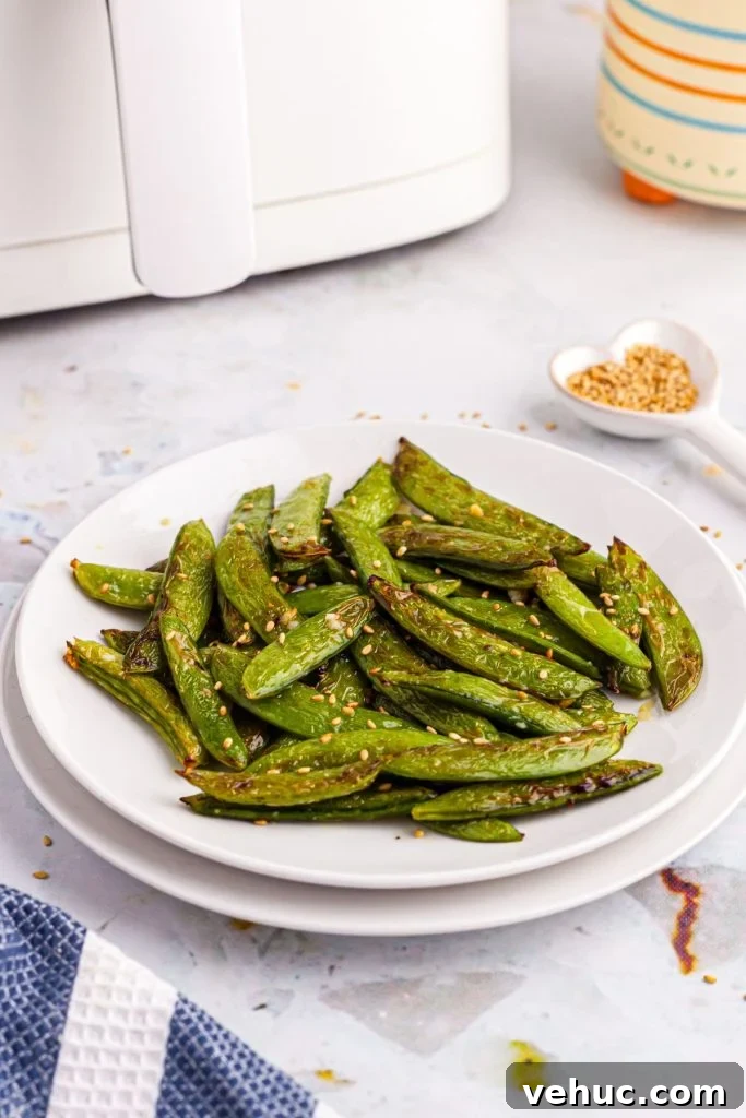 Crispy Air-Fried Sugar Snap Peas in Minutes 2 White plate with snap peas in front of an air fryer with sesame seeds scattered on the table.