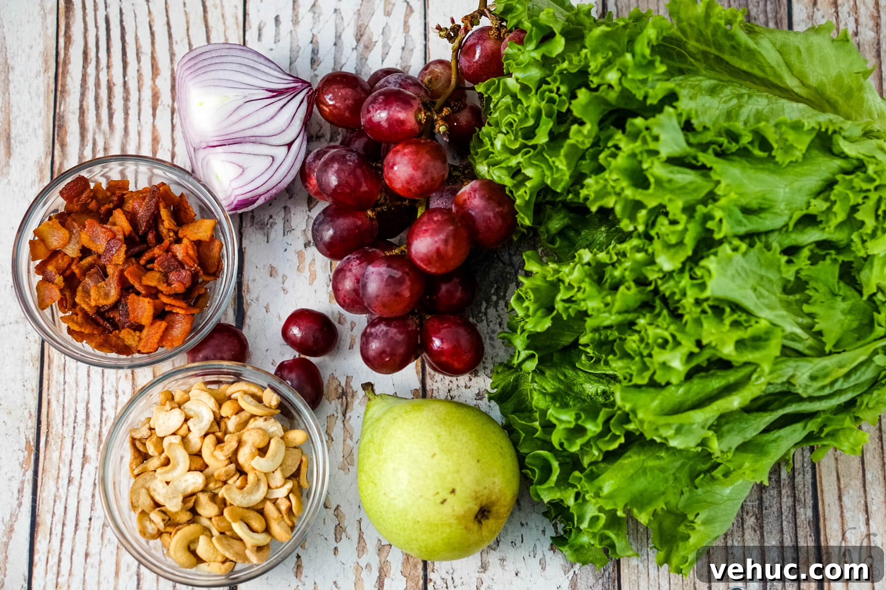 Crispy Cashew Salad with Lime Dressing: Refreshing Crunch Bowl 5 A head of lettuce, a pear, some red seedless grapes, half of a red onion, a bowl of bacon pieces and a bowl of cashew halves and pieces sit next to each other.