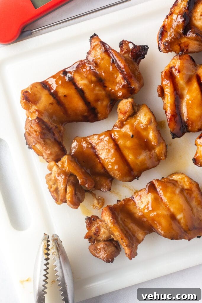 top down shot of grilled boneless skinless chicken thighs on a white cutting board with tongs and a meat thermometer next to them.