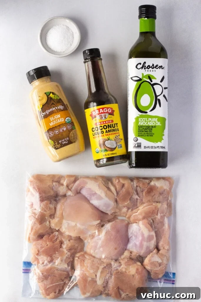 Top down shot of boneless skinless raw chicken thighs in a ziploc bag next to bottles of dijon mustard, coconut aminos, and avocado oil. A white dish with kosher salt is in the upper left corner.