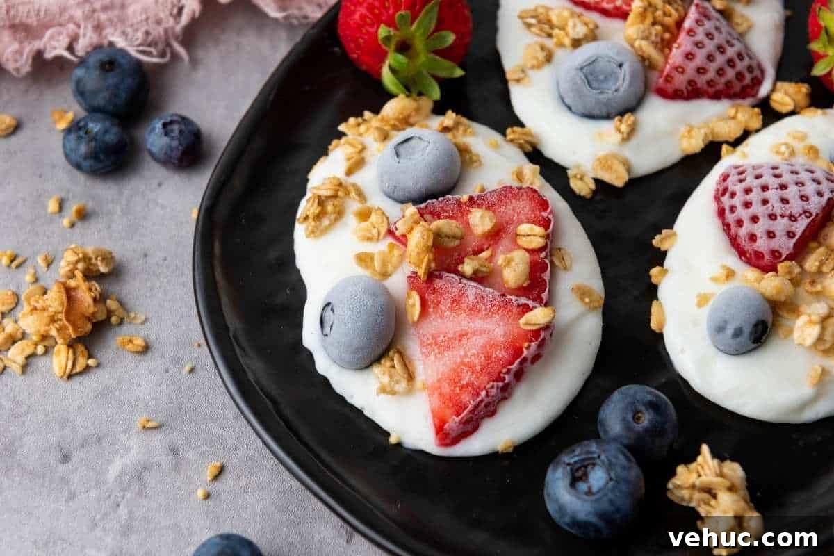 11 Strawberry Recipes That Highlight Their Sweet-Tart Flavor 7 Three Frozen Vanilla Yogurt Bites topped with sliced strawberries, blueberries, and granola are arranged on a black plate, with extra berries and granola scattered around on the gray surface.