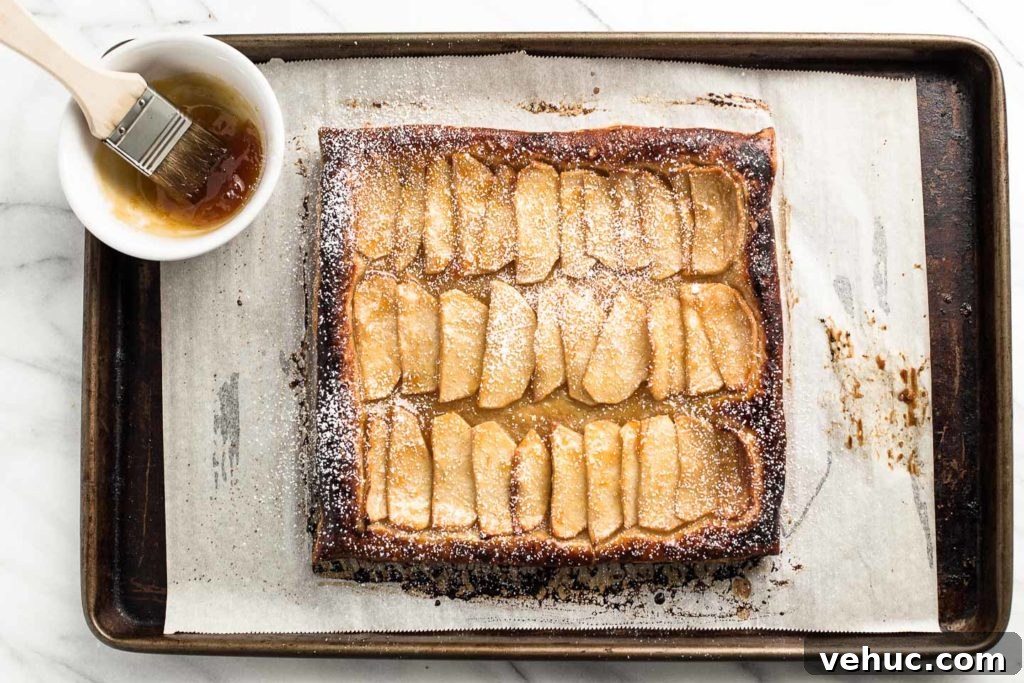 Rustic Apple Tart Recipe with Flaky Puff Pastry and Cinnamon 3 apple tart on a baking sheet with apricot jam in a bowl.