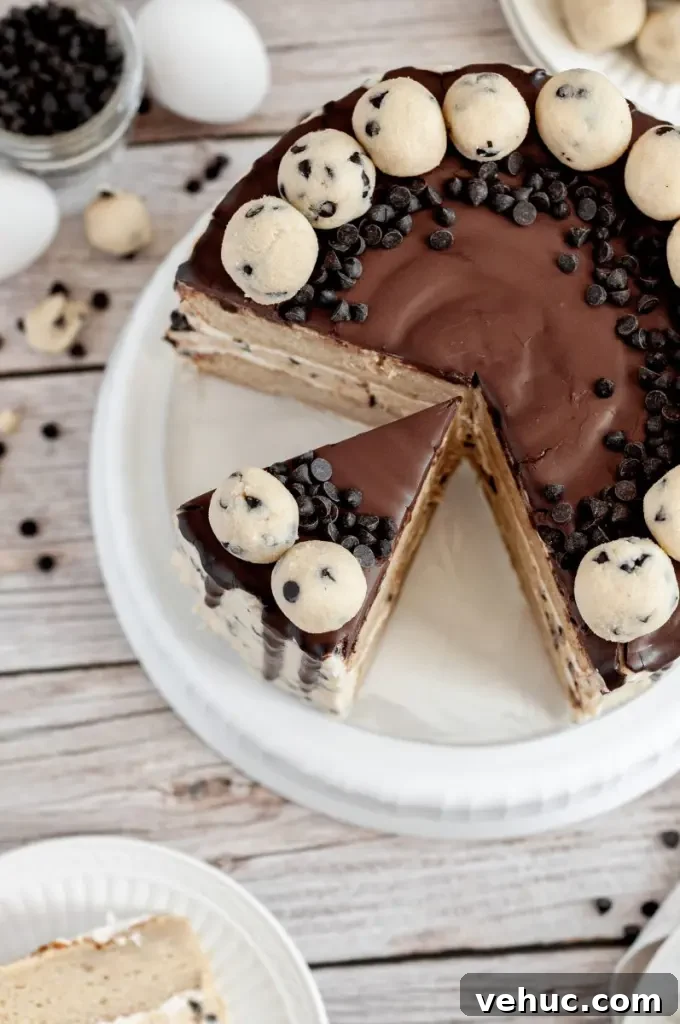 A cookie dough cake covered in cream cheese cookie dough frosting with dark brown chocolate ganache drip and topped with edible cookie dough balls.