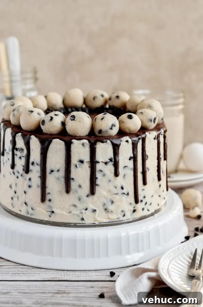 A cookie dough cake covered in cream cheese cookie dough frosting with dark brown chocolate ganache drip and topped with edible cookie dough balls.