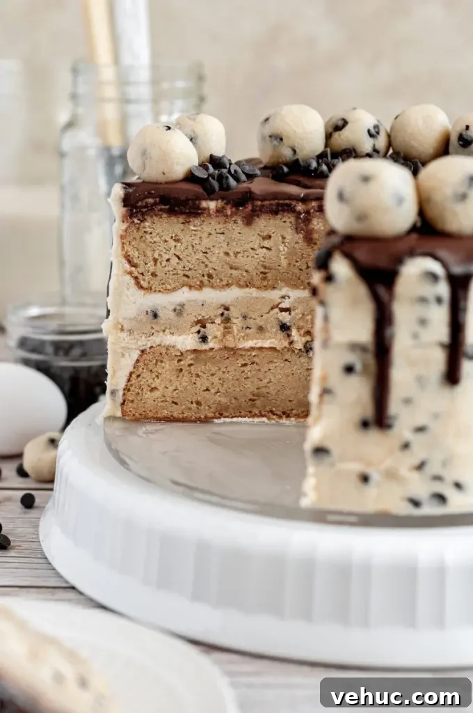 A cookie dough cake covered in cream cheese cookie dough frosting with dark brown chocolate ganache drip and topped with edible cookie dough balls.