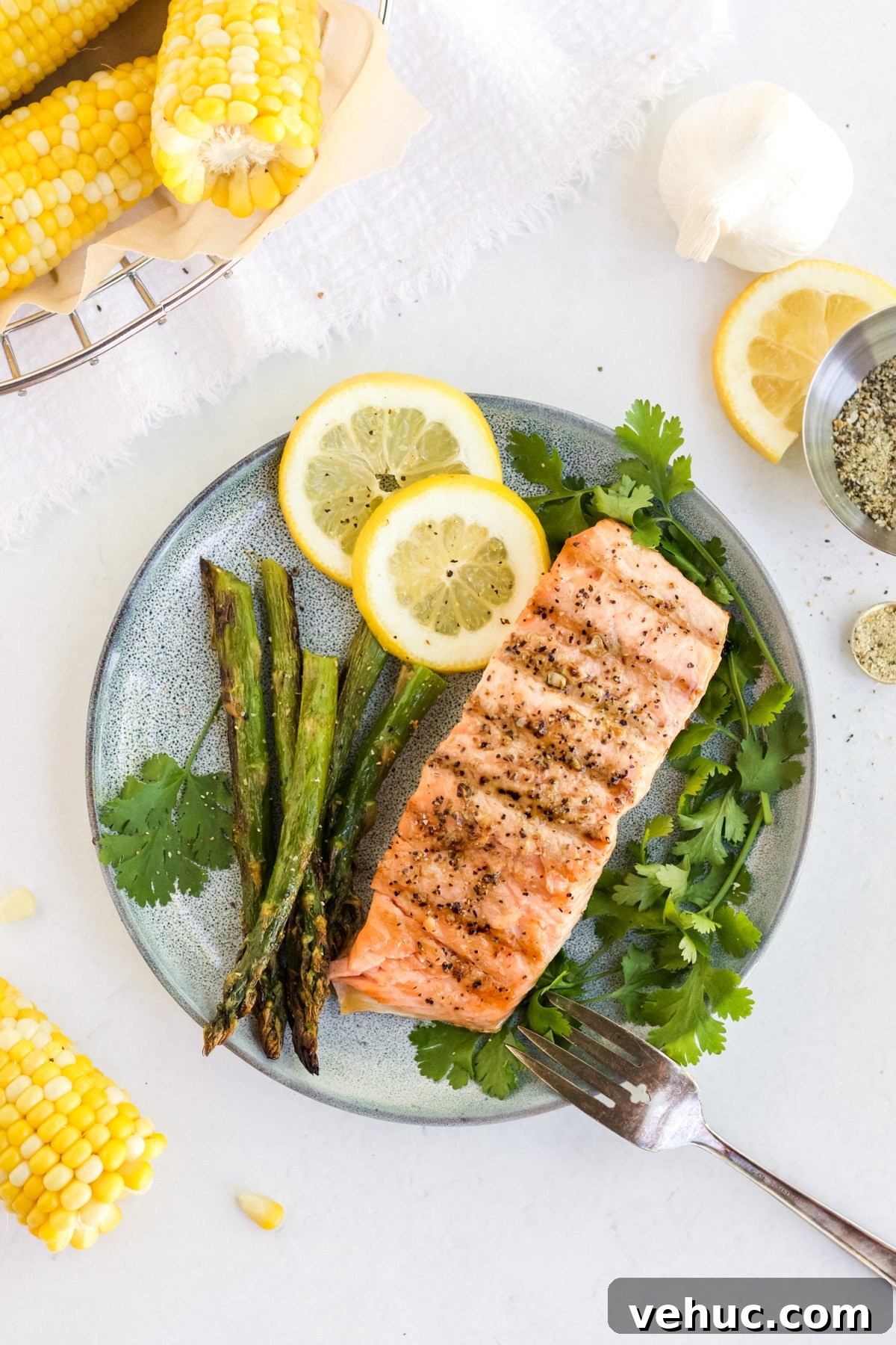 Salmon filet on a plate with herbs, asparagus and lemon, corn on the cob is off in the corner. 