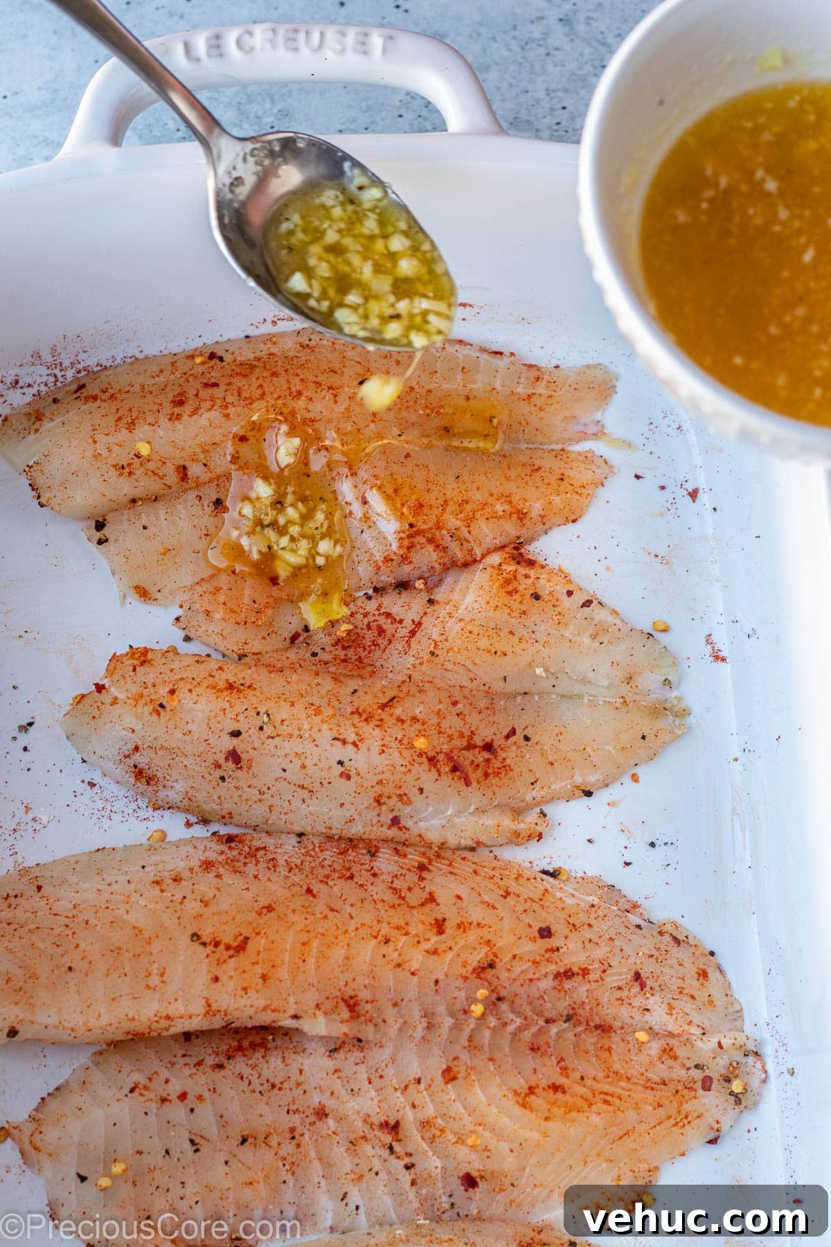 Garlic Butter Tilapia Recipe with Lemon and Herbs 6 Spoon pouring garlic butter sauce over seasoned tilapia fillets.