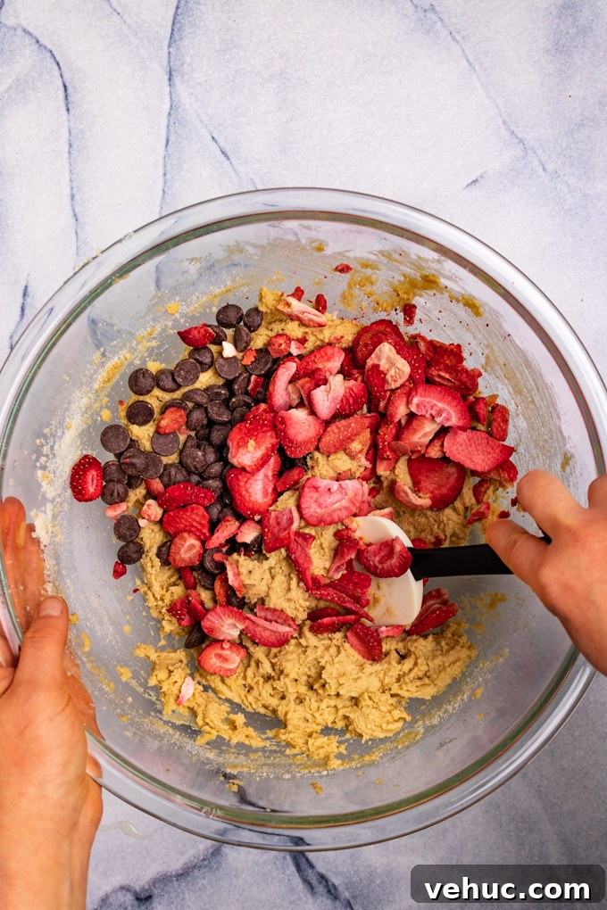 Cookie dough in a mixing bowl with freeze dried strawberries and chocolate chips being stirred in by a spatula - hands are holding the bowl.