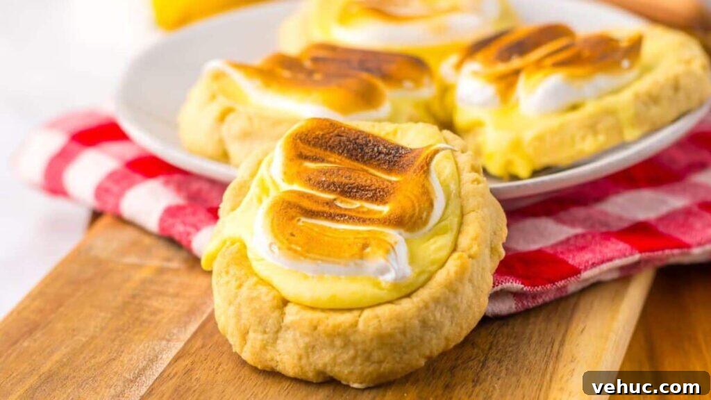 Four lemon meringue cookies with browned meringue on a plate and a wooden board, placed on a red and white checkered cloth.