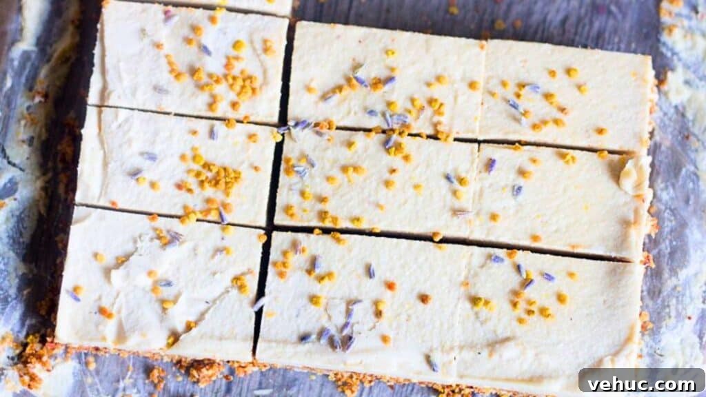 A plate with three stacked pieces of creamy dessert bars, topped with small yellow and purple sprinkles. The bars have a crumbly brown base. A fork is placed next to the bars on the patterned plate.
