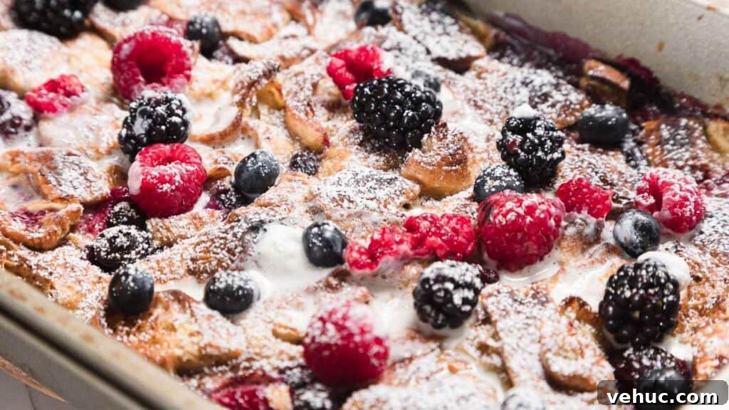 Close-up of a baked dessert topped with powdered sugar, featuring blackberries, raspberries, and blueberries. The dish is in a rectangular baking pan, with visible golden-brown edges and drizzles of cream or sauce.