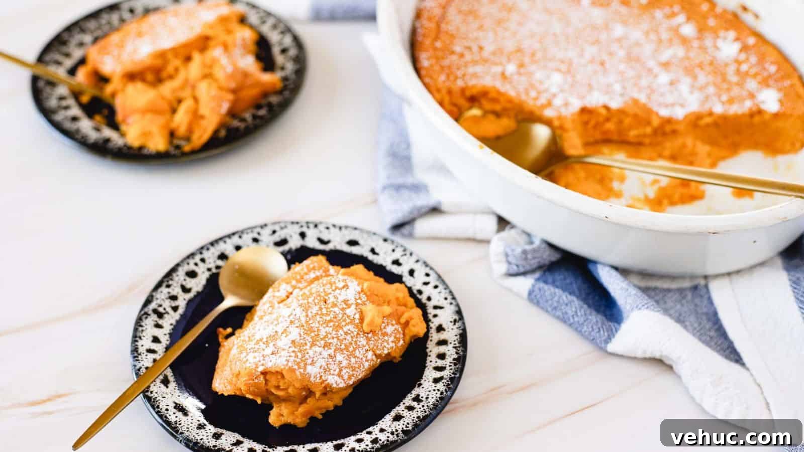 A slice of sweet potato casserole with a dusting of powdered sugar is served on a black and white plate. A gold spoon rests beside it. In the background is a casserole dish with more of the dessert and a white and blue towel.