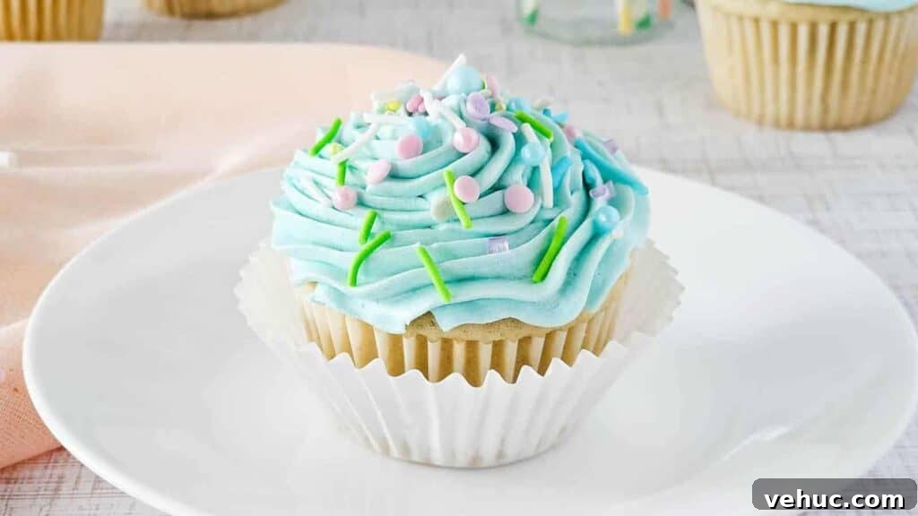 A vanilla cupcake with light blue frosting sits on a white plate. The frosting is topped with pastel sprinkles and green icing. The cupcake is in a white paper liner. A light pink cloth is partially visible in the background.