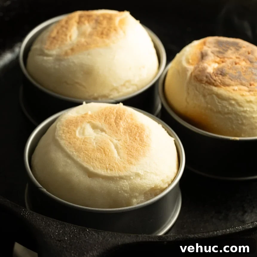 Three fully-risen English muffins in metal molds in a cast iron skillet, with tops more deeply browned and slightly uneven in color.