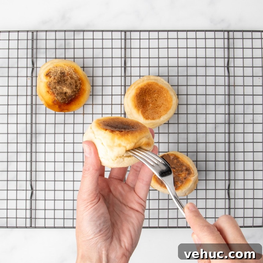 A hand using a fork to pierce the side of a browned English muffin while four cooked muffins rest on a wire rack.
