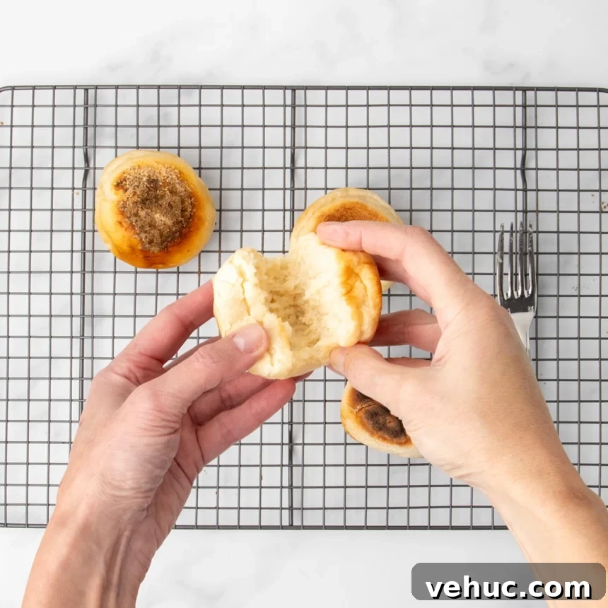 Two hands pulling apart a cooked English muffin to reveal the airy interior, with additional muffins and a fork on a wire rack in the background.