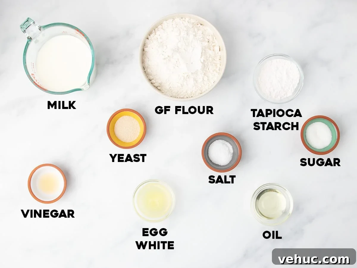 Ingredients for gluten free English muffins arranged on a white marble surface in small bowls and cups, each labeled milk, GF flour, tapioca starch, sugar, salt, yeast, vinegar, egg white, and oil.