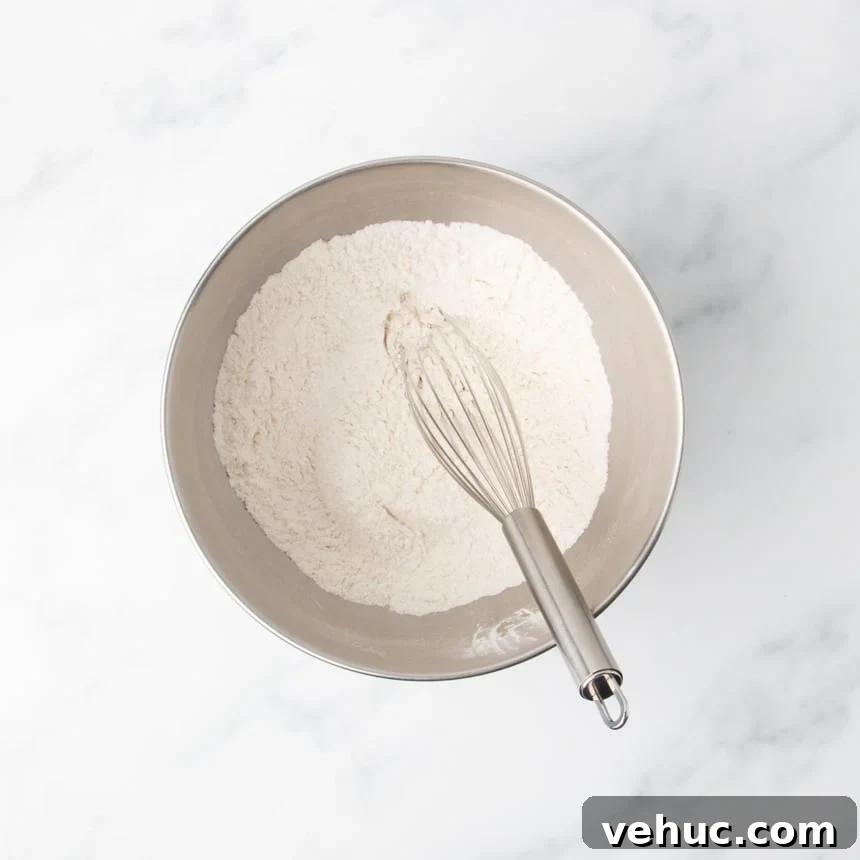 A metal mixing bowl filled with a dry flour mixture with a metal whisk resting inside on a white marble surface.