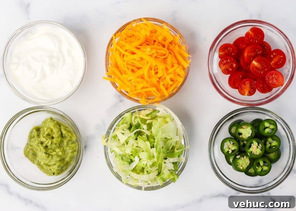 High-Protein Keto Taco Bowls for Low-Carb Meal Prep 6 Toppings ready to be assembled for keto taco bowls including lettuce, sliced cherry tomatoes, shredded cheddar cheese, guacamole, sour cream, and sliced jalapenos