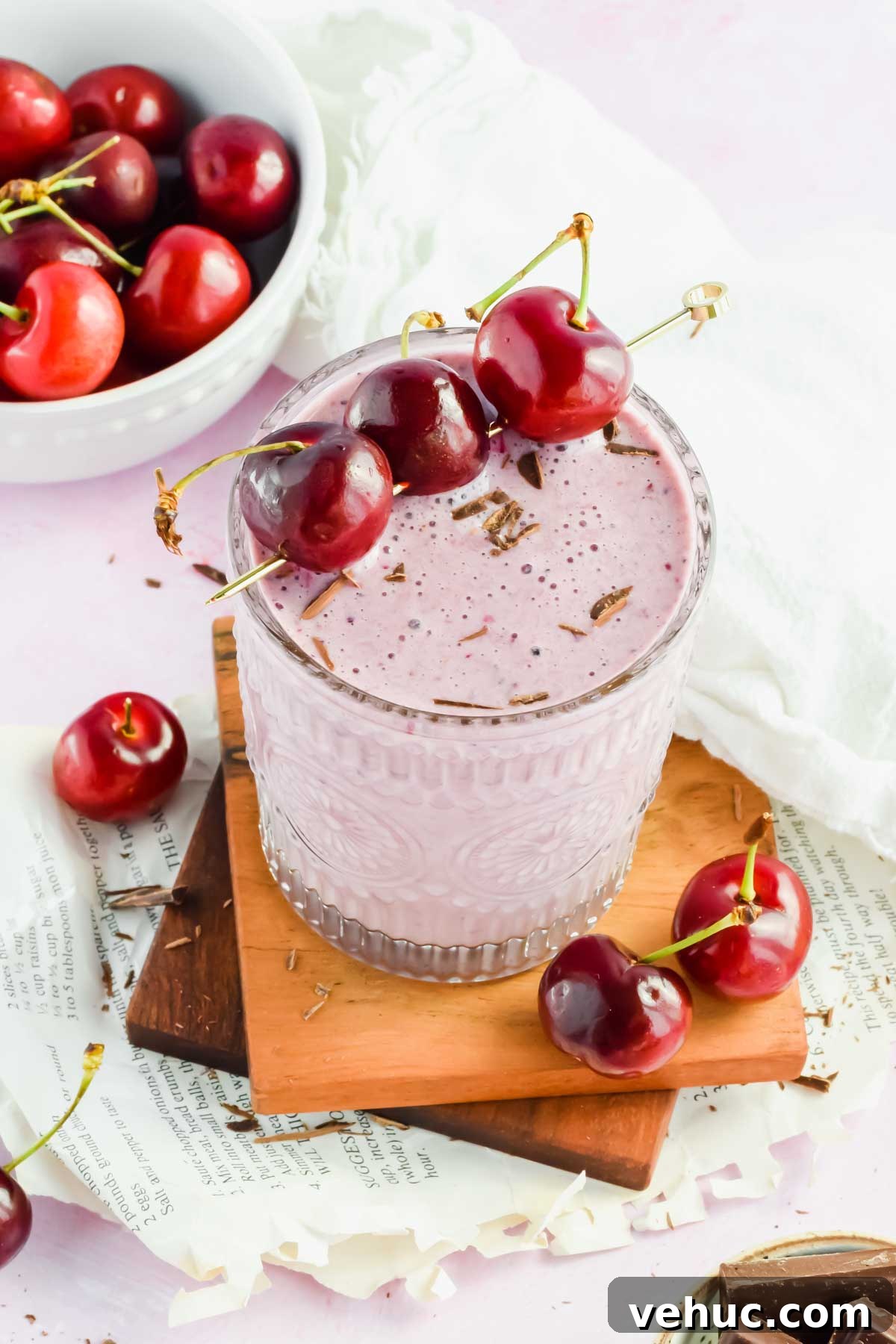 clear glass filled with light pink chocolate cherry smoothie garnished with fresh cherries on a skewer laying across top of glass on wood block with white background and cherries scattered around.