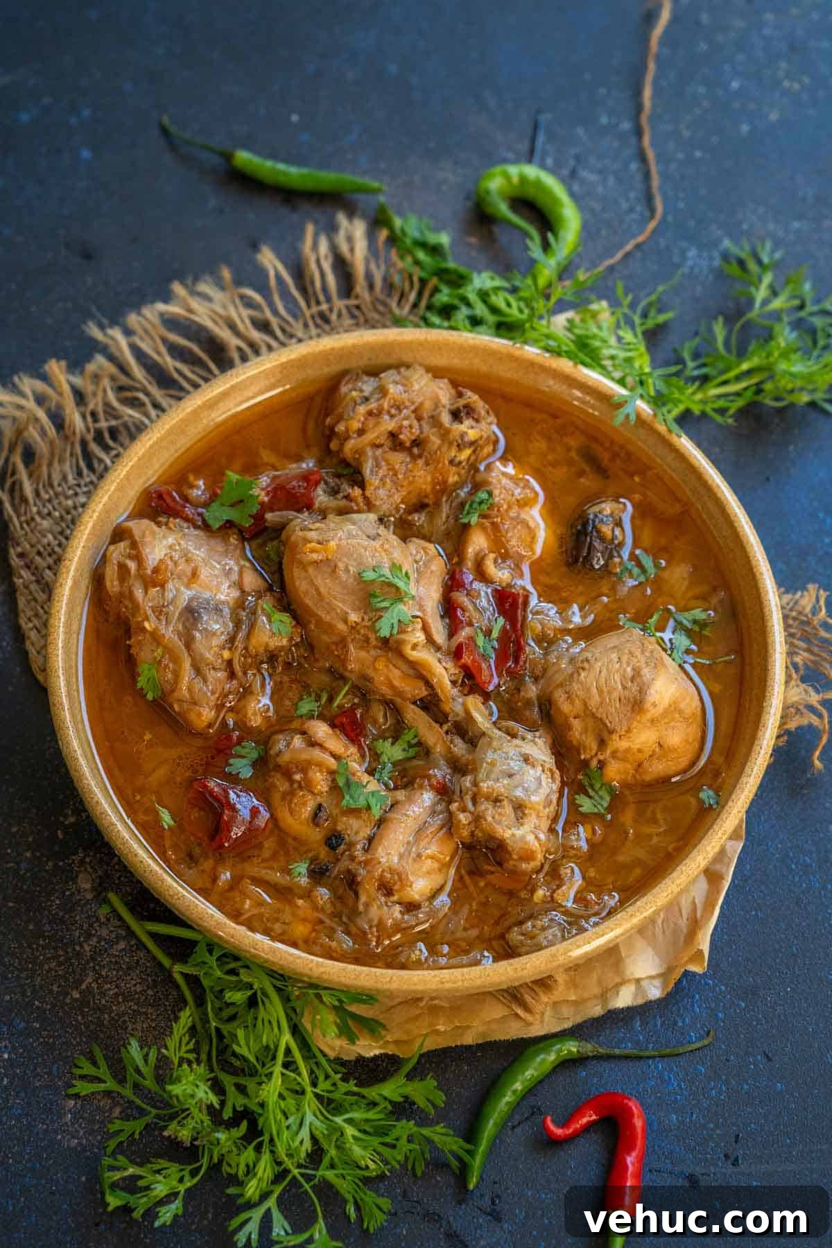 Mughlai Chicken Stew Recipe: Aromatic Creamy Indian-Style Stew 2 A bowl of spicy Mughlai Chicken Stew garnished with fresh cilantro sits on a rustic cloth, surrounded by red and green chilies and sprigs of cilantro on a dark surface.