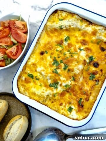 A white enamel baking dish filled with cheese-toppped lasagne. There is a dish of tomato salad in the background and two crusty rolls on a plate.