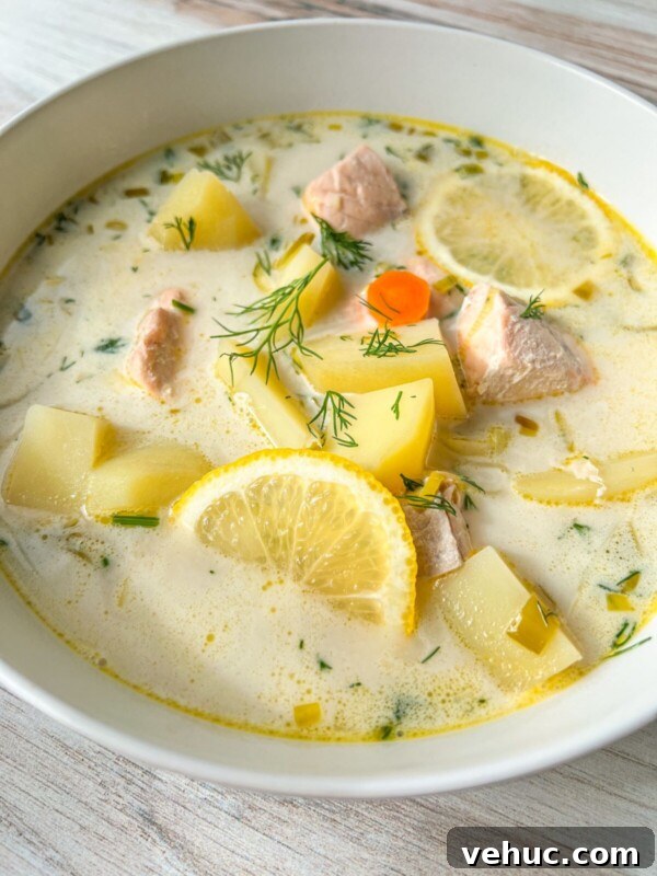 Authentic Finnish Salmon Soup Recipe (Lohikeitto) 7 A bowl of creamy Finnish Salmon Soup with chunks of salmon, potatoes, carrot slices, lemon wedges, and fresh dill, served in a white bowl on a light wooden surface.