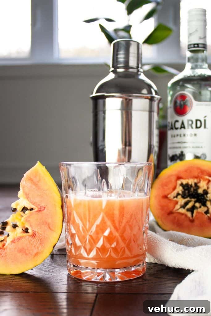 Glass of papaya rum punch beside half a papaya and a papaya slice with a rum bottle and cocktail shaker in the background