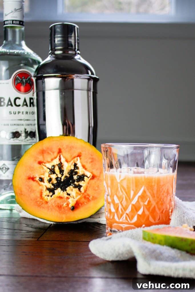 Glass of papaya rum punch beside half a papaya with a rum bottle and cocktail shaker in the background