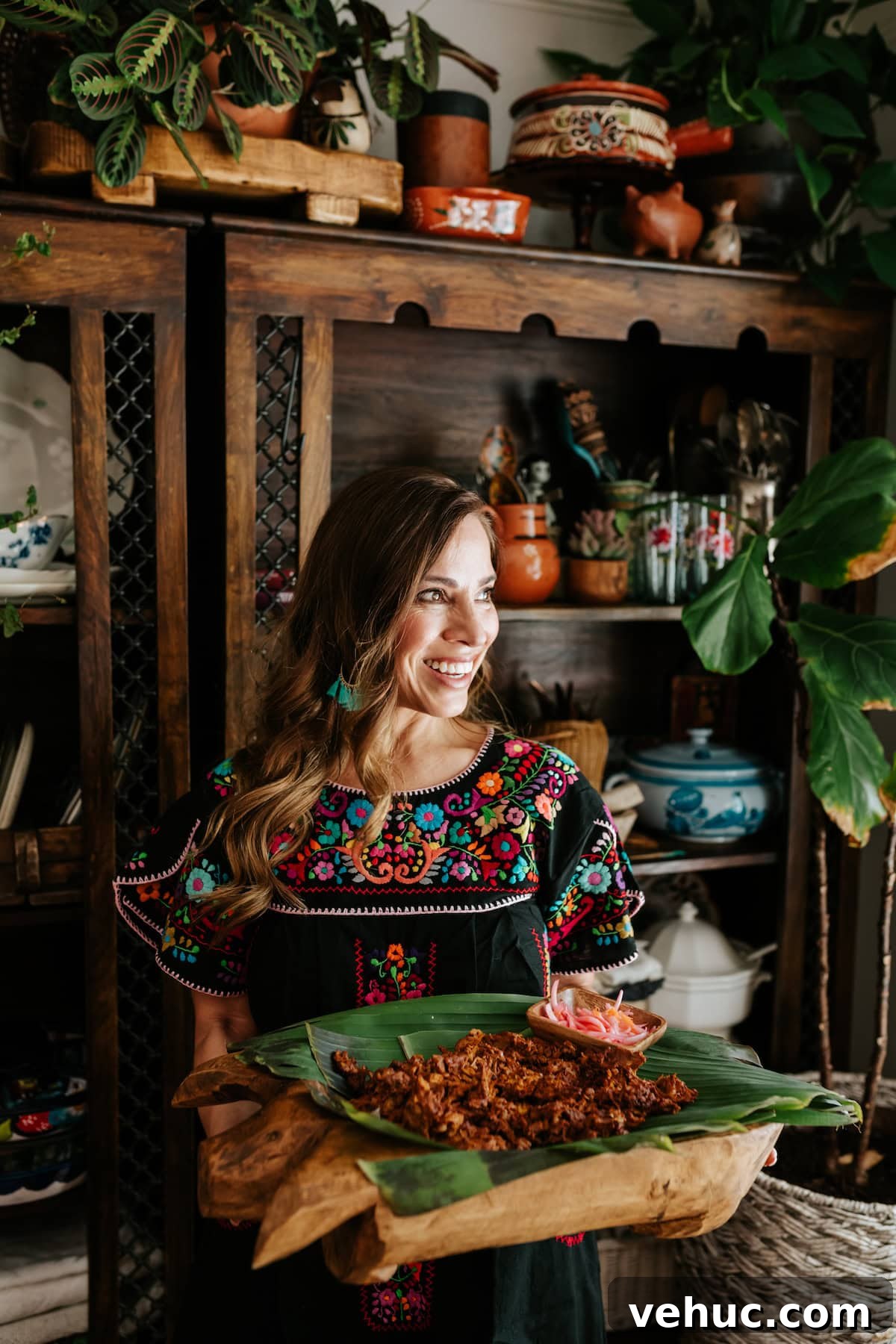 Authentic Cochinita Pibil Tacos Recipe — Yucatan Slow-Roasted Pork 8 Yvette Marquez holding a wooden platter of Cochinita Pibil with pickled onions, wearing a black embroidered dress in a Mexican-inspired kitchen.