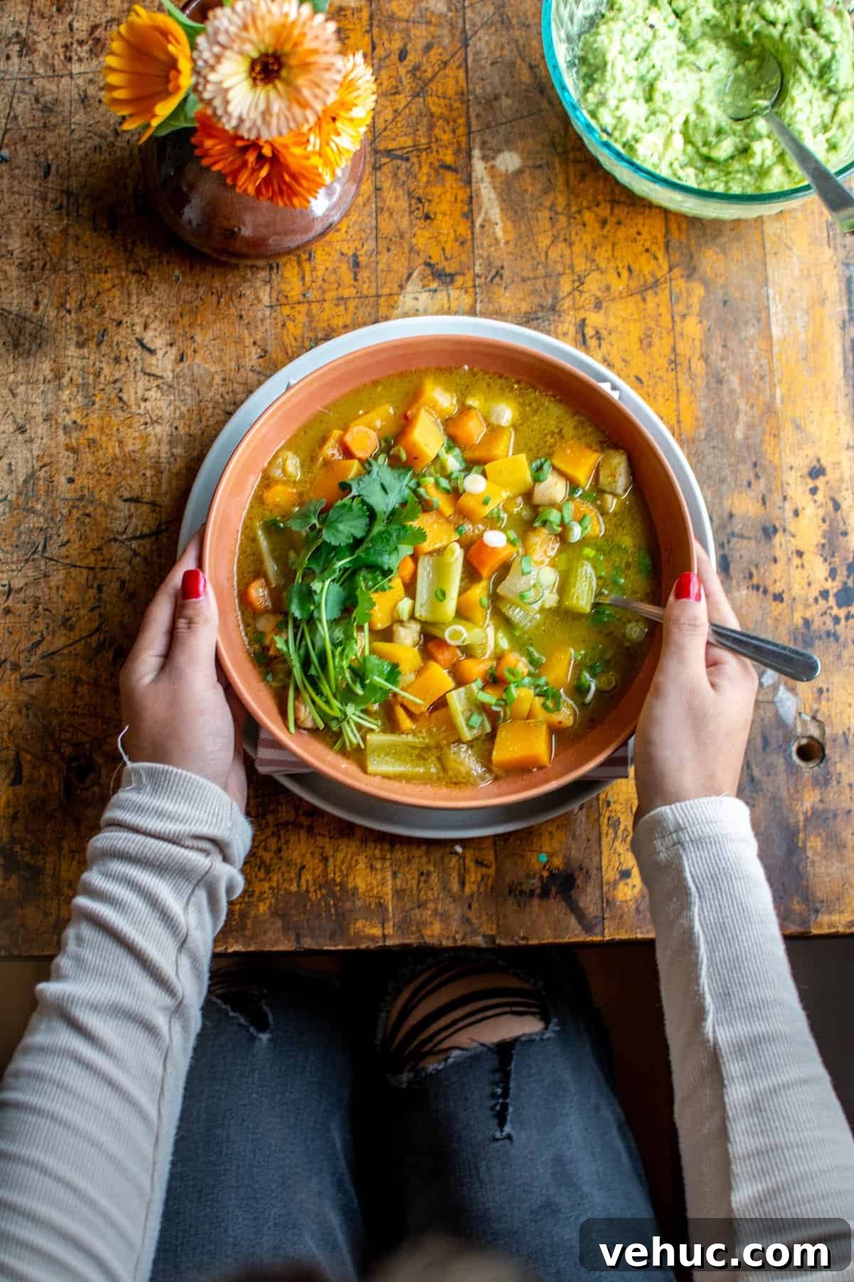 Tangy Chicken and Tomatillo Stew Recipe for Weeknight Dinners 5 A woman holding a bowl of chicken tomatillo stew sitting at a wood table with a vase of flowers next to it.