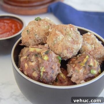 A bowl of homemade chicken meatballs with visible herbs, accompanied by a side of tomato sauce on a marble countertop.