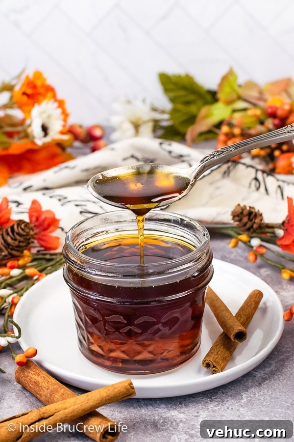 Cinnamon Maple Syrup drizzling A clear jar on a white plate filled with cinnamon syrup with a spoon drizzling more syrup into it.