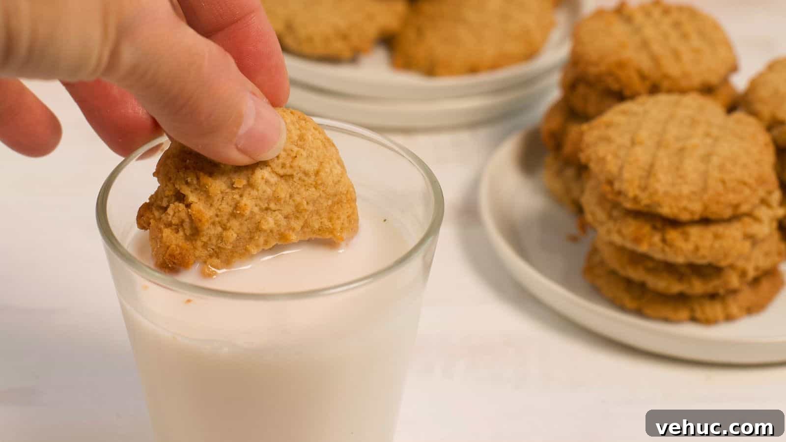 A hand dipping a peanut butter cookie into a glass of milk, with more cookies stacked on a plate in the background.