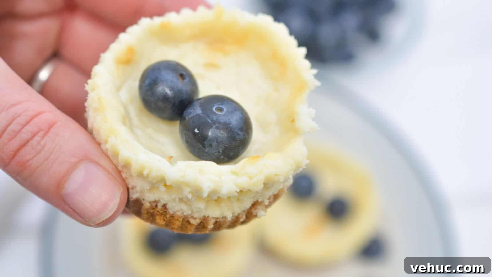 A hand holding a mini cheesecake topped with two blueberries, with more cheesecakes and blueberries on a plate in the background.