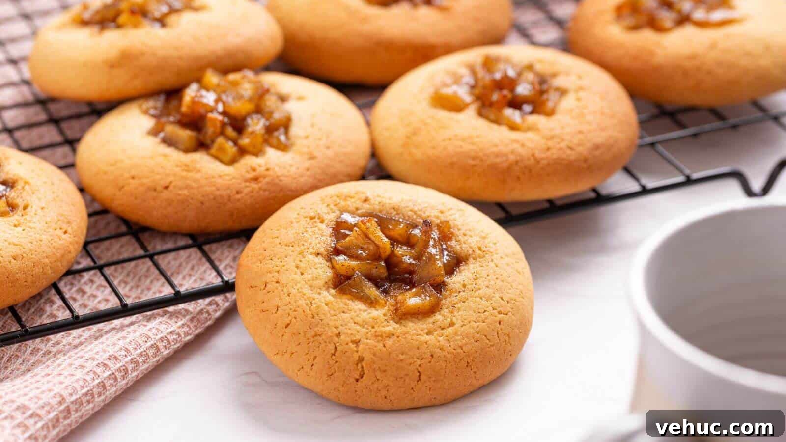 Freshly baked apple pie cookies topped with caramelized fruit on a cooling rack, with a cup and a cloth nearby on a light colored surface.