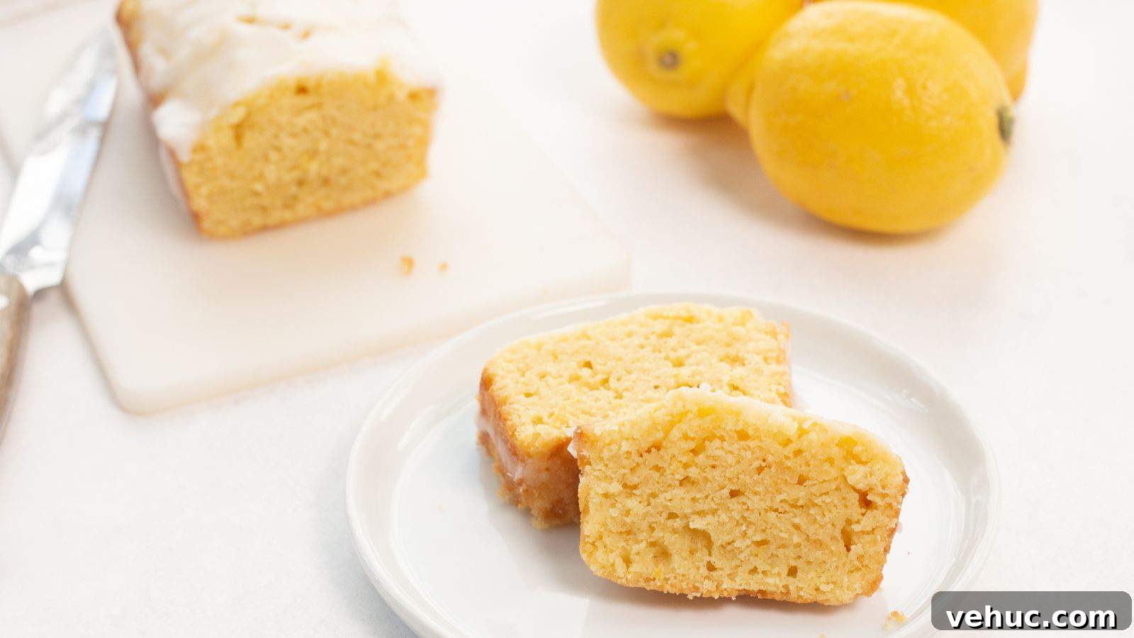 Slices of lemon loaf with icing on a white plate, with whole lemons in the background.