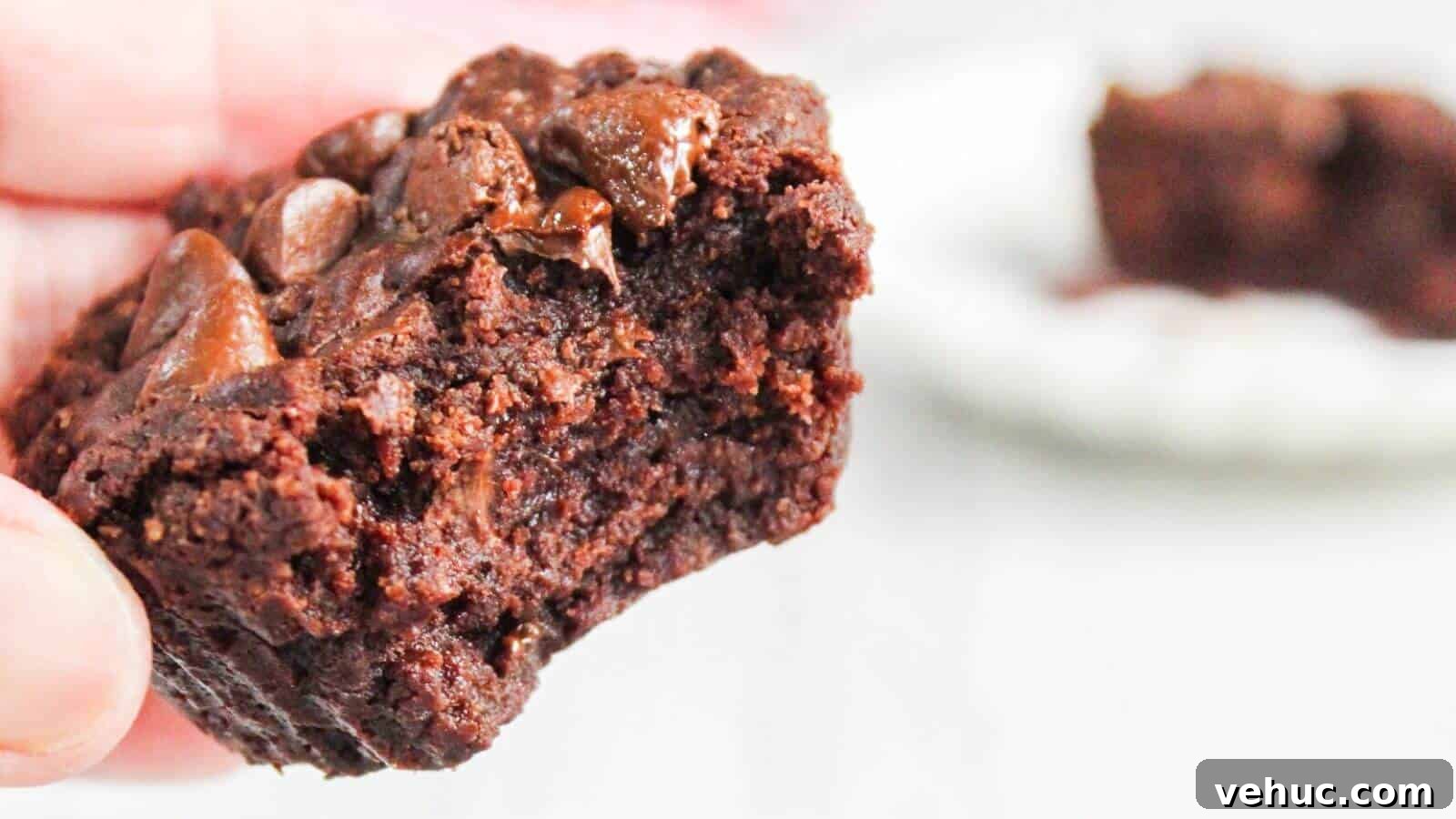 A close-up of a hand holding a half-eaten chocolate brownie with visible melted chocolate chips.