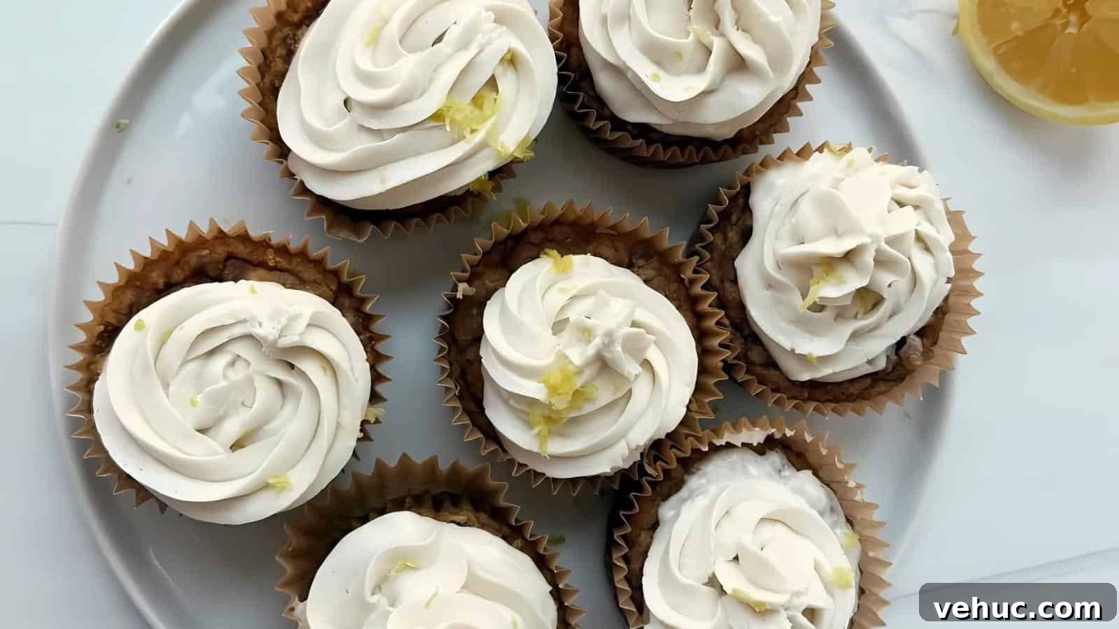 Seven cupcakes with white frosting and lemon zest are arranged on a white plate; a lemon slice is partially visible in the top right corner.