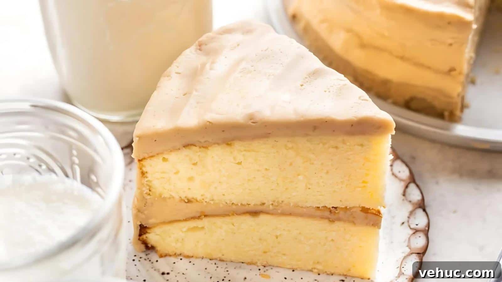 A slice of yellow layer cake with light brown frosting sits on a plate, with a glass of milk and part of the remaining cake visible in the background.