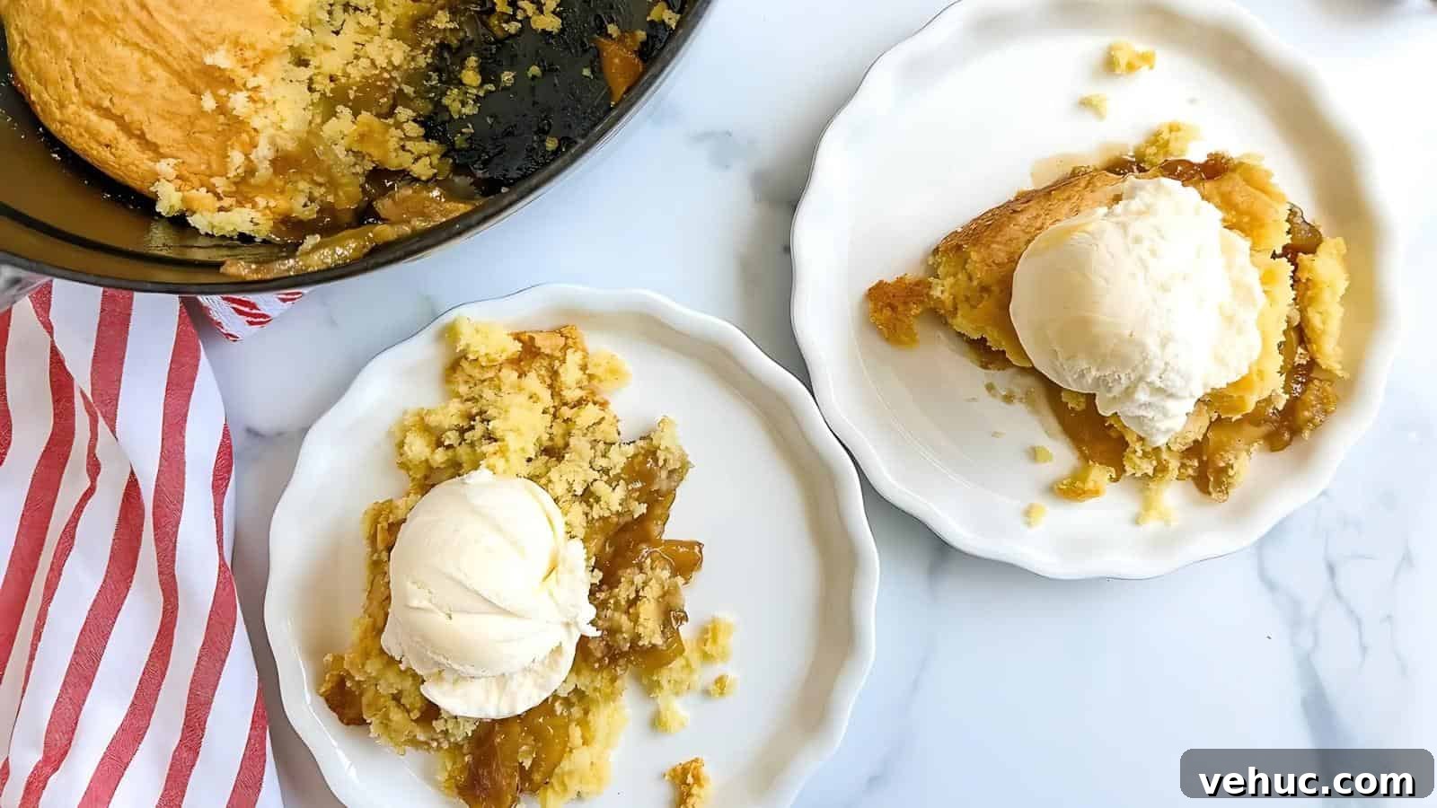 Two white plates with servings of fruit cobbler topped with vanilla ice cream, next to a baking dish with remaining cobbler and a red-striped napkin on a marble surface.