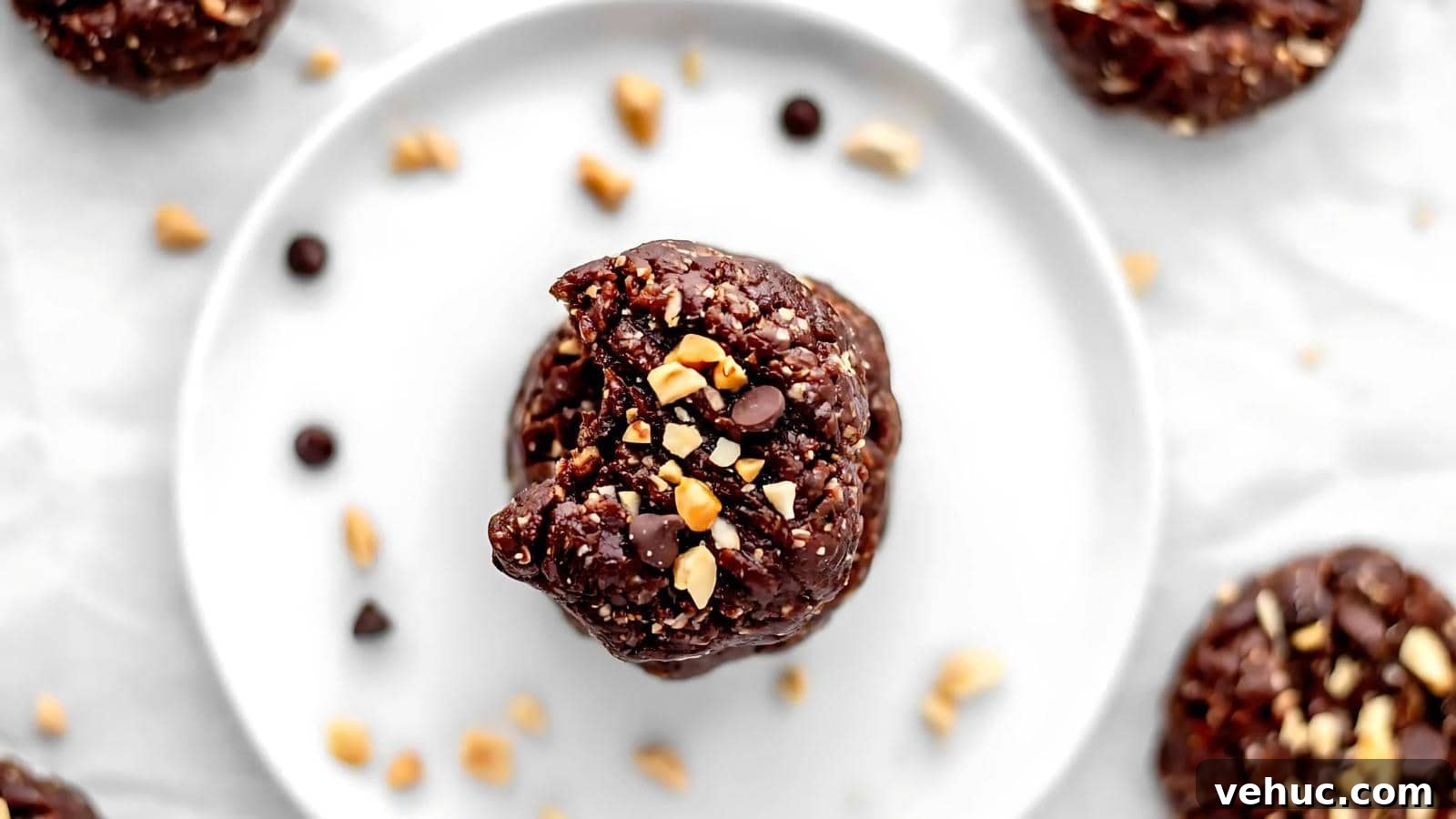 A close-up of a chocolate cookie topped with chopped peanuts and chocolate chips on a white plate, surrounded by more cookies and scattered toppings.
