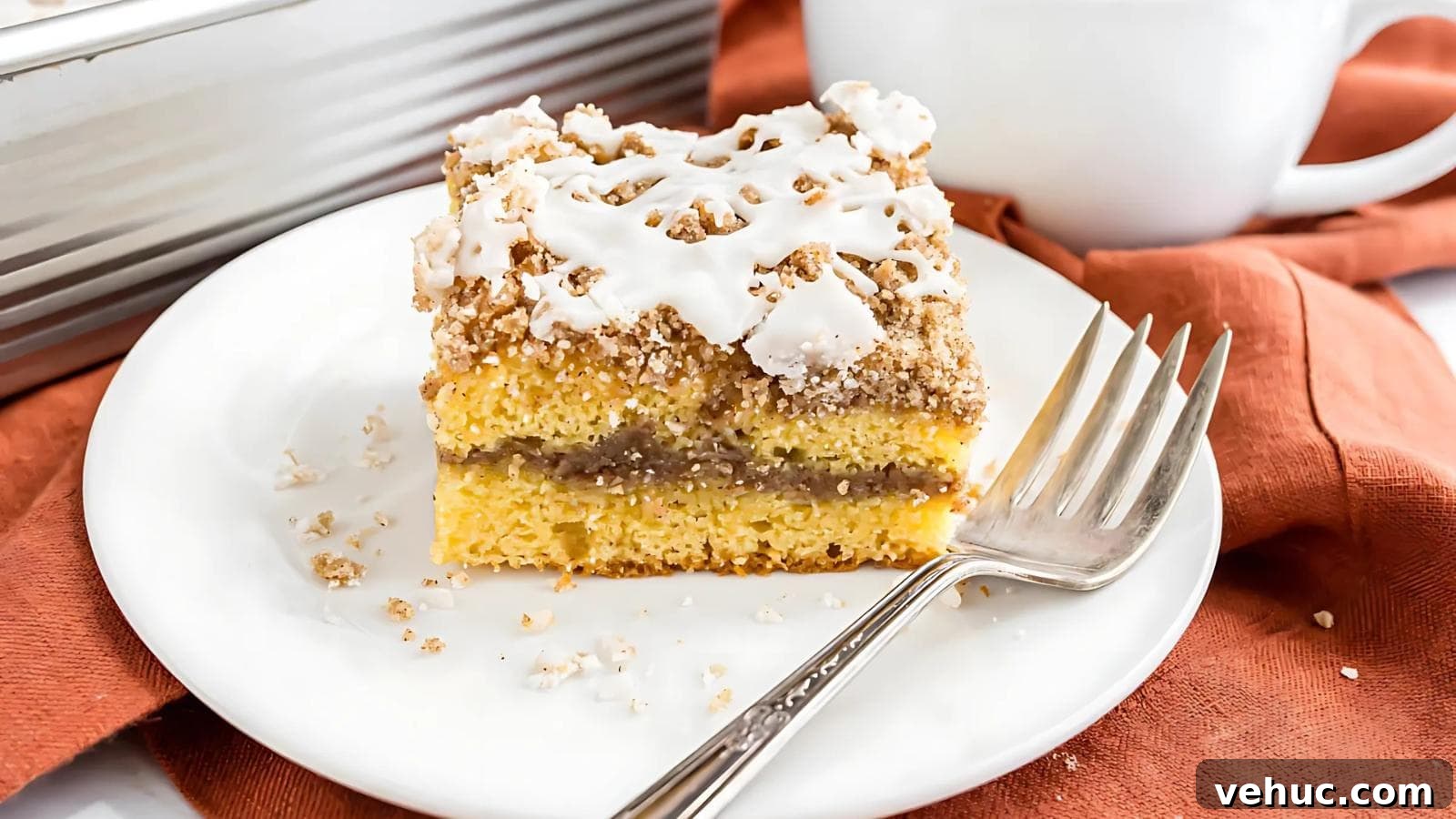 A slice of coffee cake with icing and crumb topping sits on a white plate with a fork, on an orange napkin.