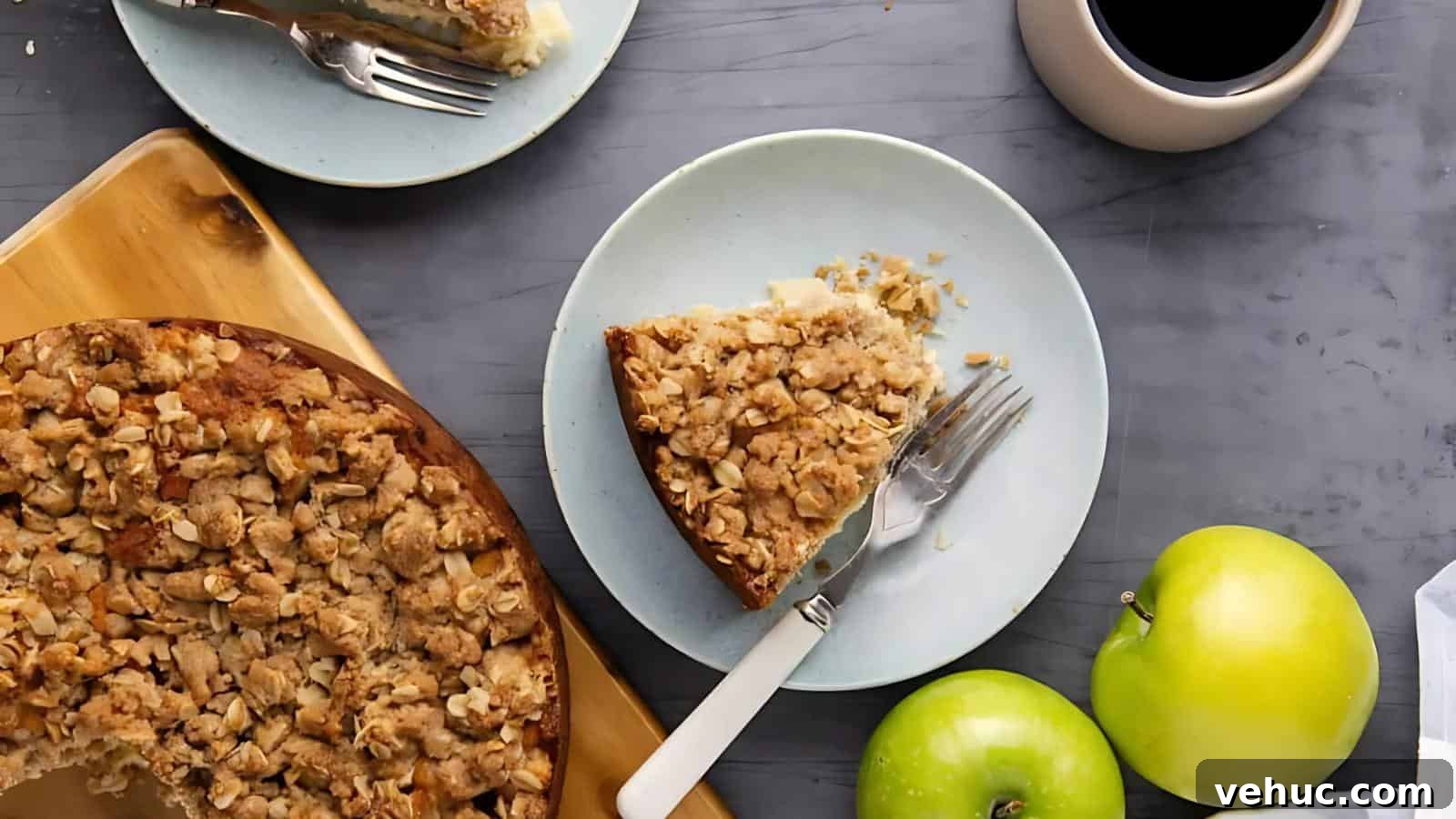 A slice of crumb-topped apple cake on a plate with a fork and knife, next to the whole cake, two green apples, and a cup of coffee on a gray surface.