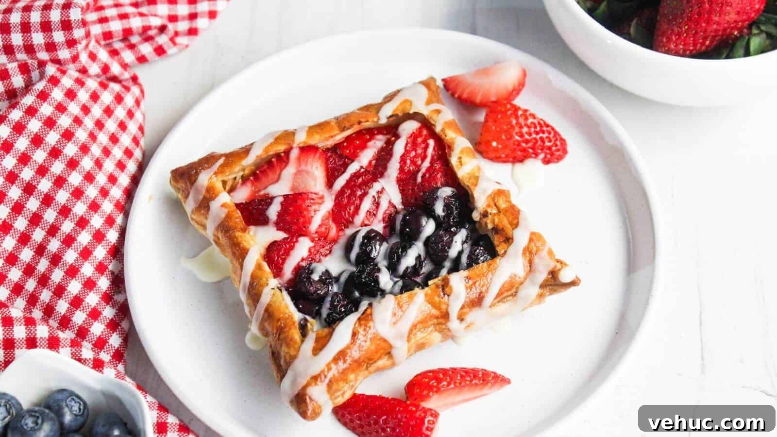 A cream cheese danish topped with strawberries and blueberries drizzled with icing is on a white plate. A red checkered cloth, a bowl of strawberries, and a small bowl of blueberries are nearby.
