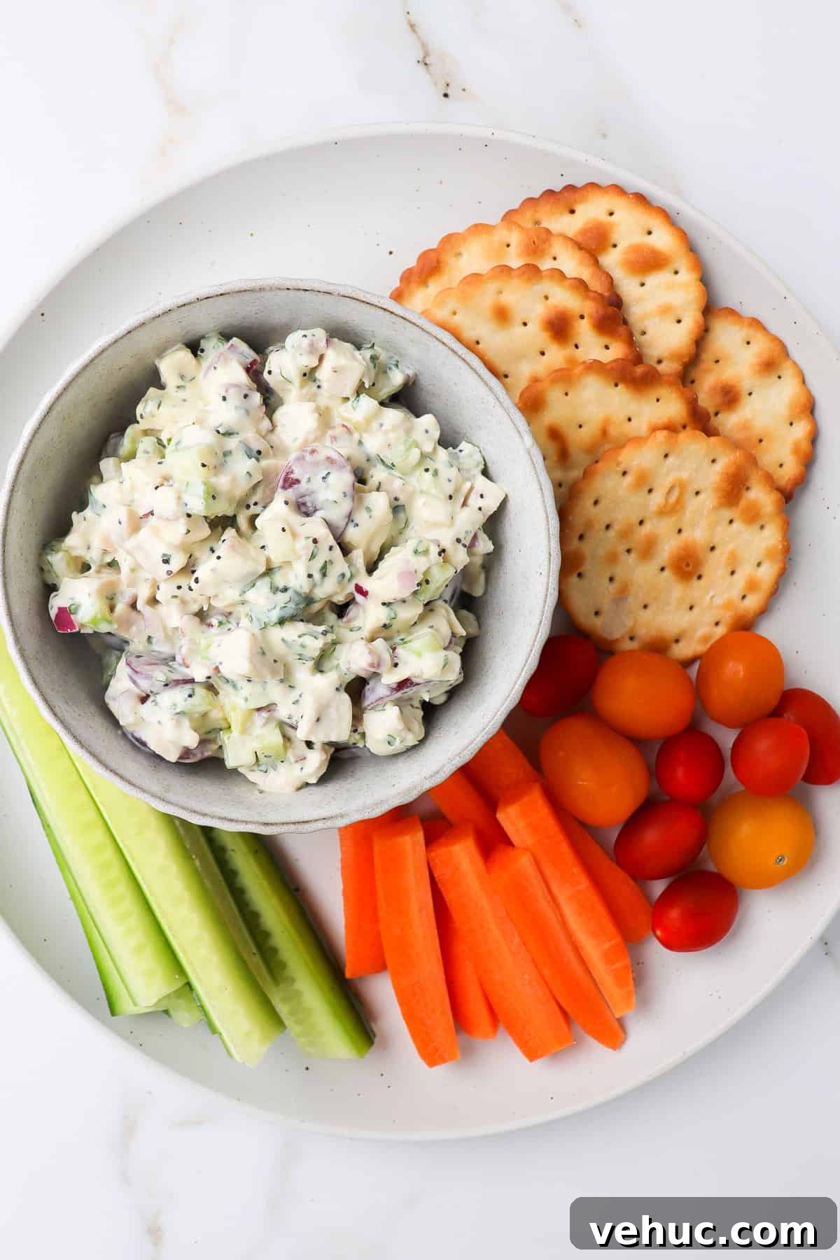 Mixed plate with chicken salad in a bowl, carrot sticks, cucumbers, grapes and crackers on the side.