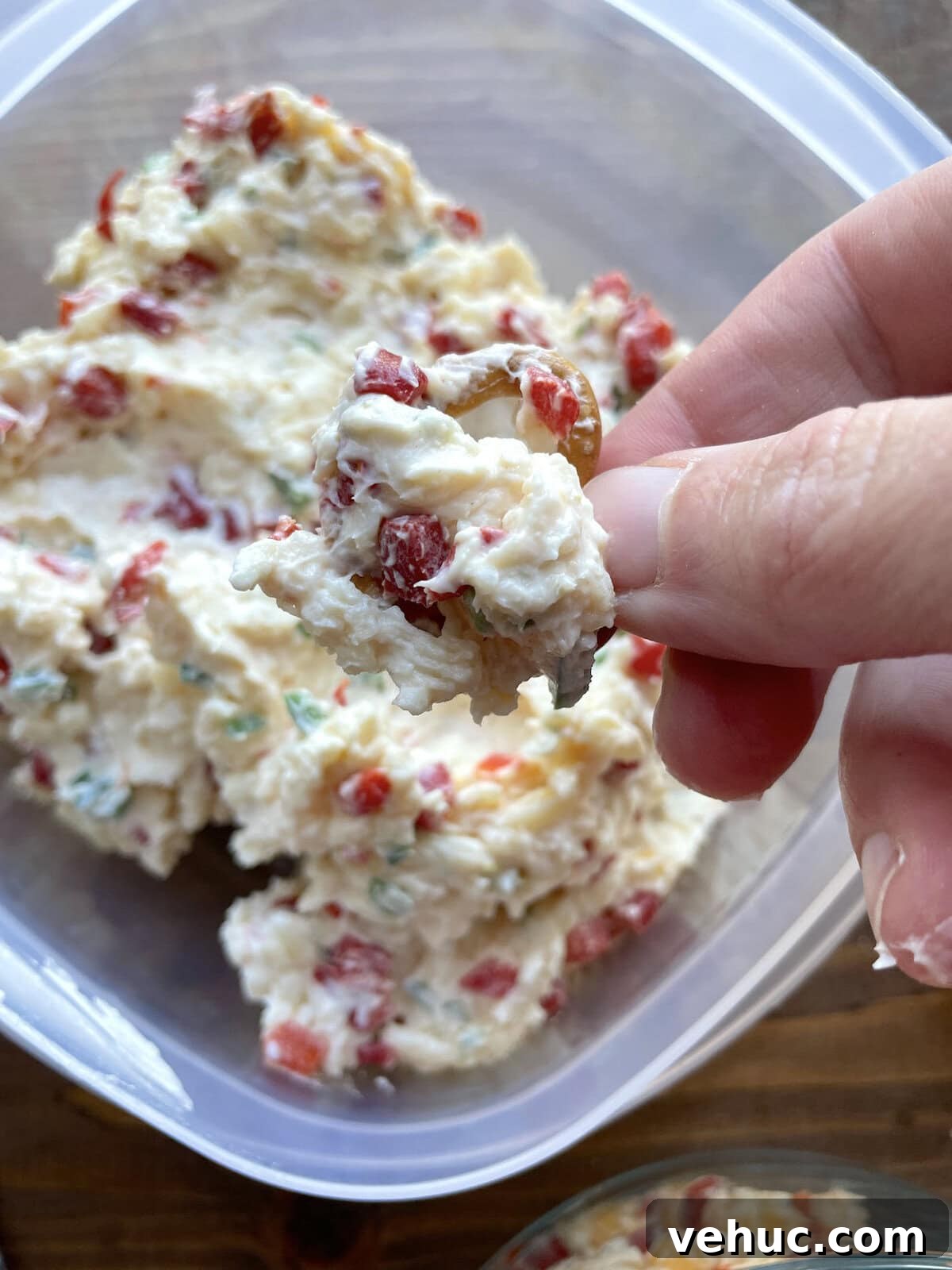 Spicy Jalapeño Pimento Cheese Dip Recipe for Parties 6 A hand holding scoop of pimento cheese with jalapeños on a pretzel over a Tupperware.