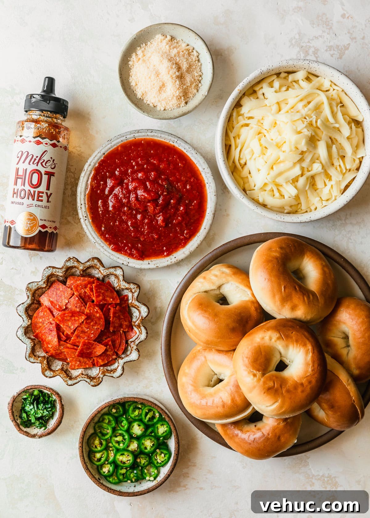 Homemade Pizza Bagel Bites Recipe: Quick Oven & Air Fryer Guide 3 White and brown bowls of bagels, tomato sauce, mozzarella, parmesan, pepperoni, basil, and serrano peppers next to a bottle of hot honey on a tan counter.