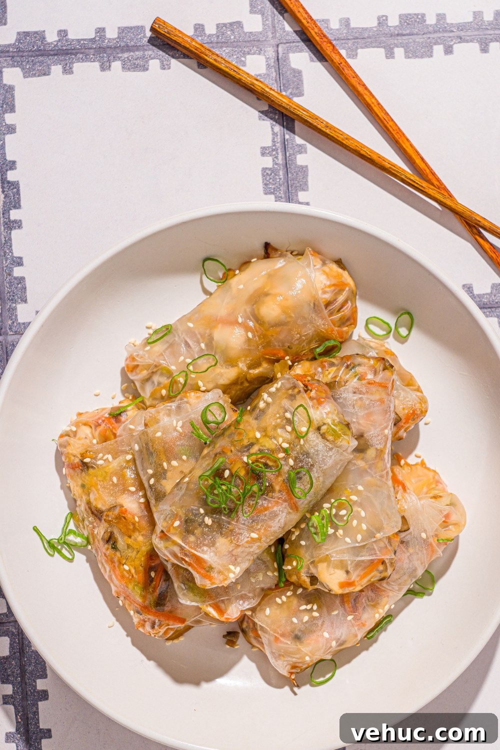 Crispy Rice Paper Veggie Dumplings: Viral TikTok Recipe 11 A white bowl with vegetable rice paper dumplings garnished with sliced green onions and sesame seeds. The dumplings are lightly fried. The bowl is on a tiled counter with a pair of chopsticks and small.