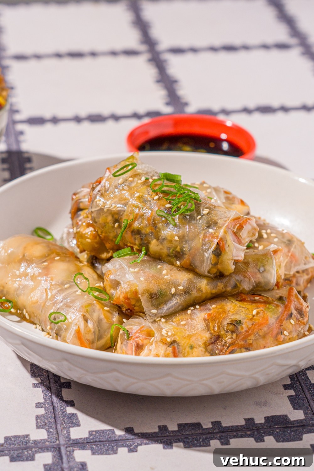 Crispy Rice Paper Veggie Dumplings: Viral TikTok Recipe 3 A white bowl with vegetable rice paper dumplings garnished with sliced green onions and sesame seeds. The dumplings are lightly fried. The bowl is on a tiled counter with a pair of chopsticks and small bowl of dipping sauce.